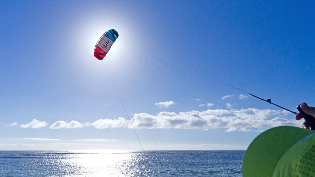 A large blue, white and red kite being flown in the air over the sea with a person in the bottom right corner in a green cap holding the kite strings.