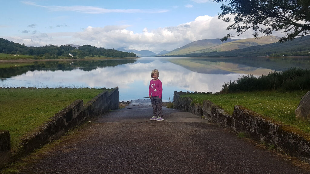 Peter's daughter, a blonde haired little girl, stands on the side of a lake with green hills in the distance on their first post-lockdown family motorhome holiday
